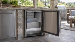 A stainless steel outdoor beverage center built into an outdoor kitchen island, door open, empty ice bin visible inside