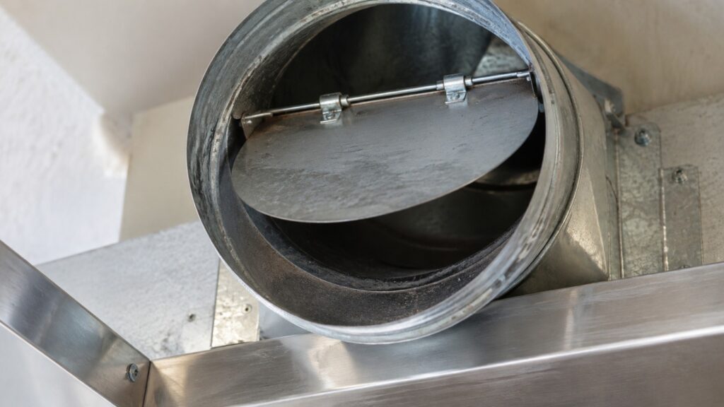 A close-up of an open range hood duct showing a hinged backdraft damper flap partway open