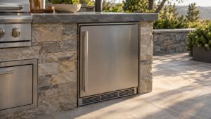 A stainless steel outdoor refrigerator built into an outdoor kitchen island in direct afternoon sun, door closed, gasket line visible around the frame