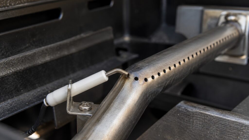 A close view of a gas grill burner with a small white ceramic electrode tip positioned a fraction of an inch from the burner port