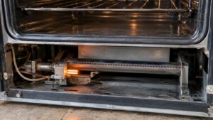 The open lower cavity of a gas oven with a small ceramic igniter glowing orange near the burner tube