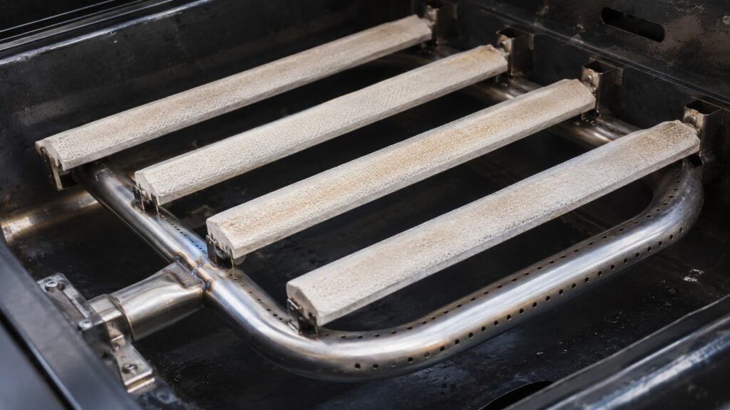 A close view of a grill burner with a U-shaped tube and ceramic radiant rods above it, no text or labels