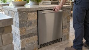 A stainless steel built-in outdoor ice maker under a counter with a hand resting near the door, no text or labels